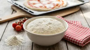 Impasto per pizza lievitato in una ciotola con farina, pomodori e pizza al forno sullo sfondo