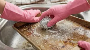 Mani con guanti rosa puliscono una teglia incrostata con una pallina di alluminio e sale nel lavello