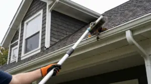 Persona pulisce una grondaia intasata con una spazzola telescopica da terra, senza salire sul tetto