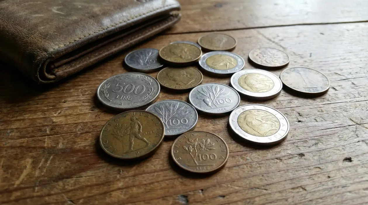 Vecchie monete italiane sparse su un tavolo di legno accanto a un portafoglio in pelle