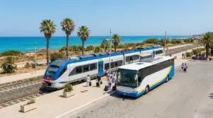 Stazione ferroviaria vicino al mare con un treno e un autobus, persone con valigie sul marciapiede