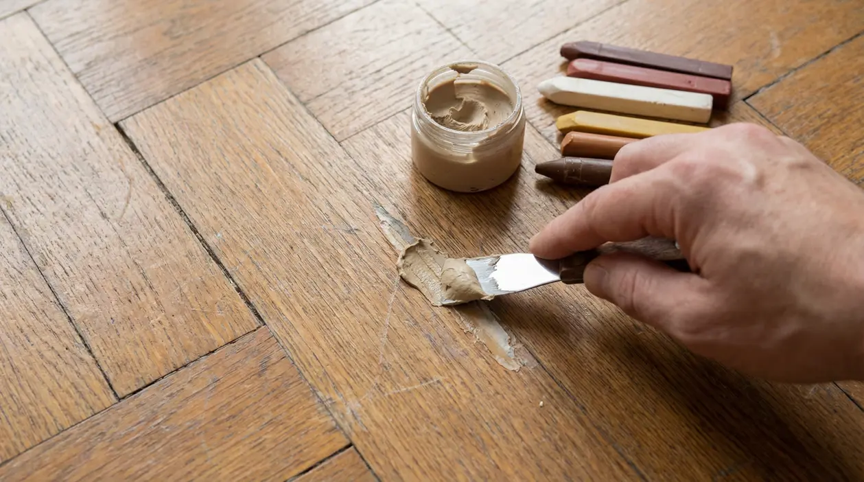 Mano che applica stucco color legno su parquet graffiato con una spatola, con cera e bastoncini ritocco accanto